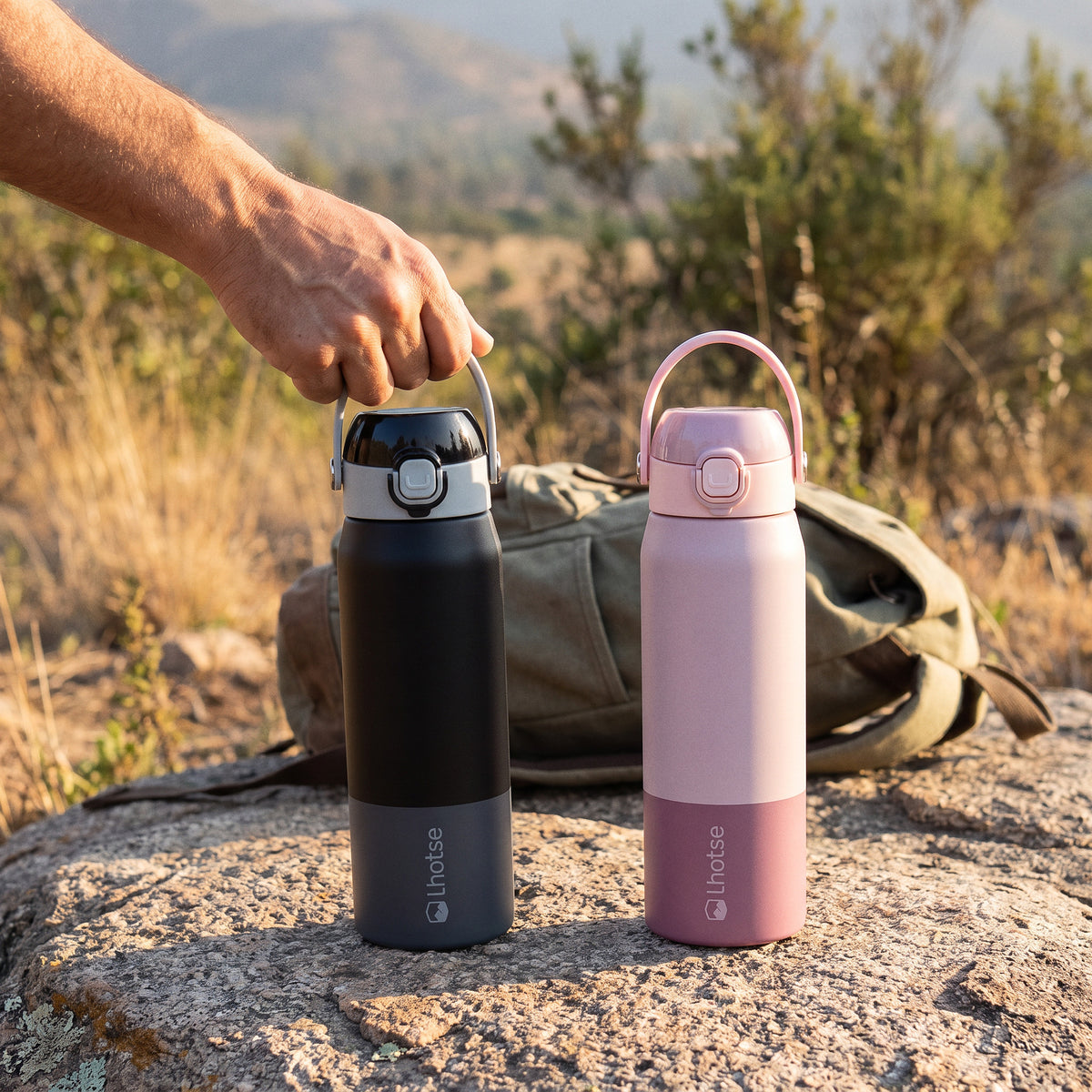Pack de 2 botellas térmicas Hydrotek 750 ml Lhotse, negra y rosa, sobre una roca en un sendero de cerro con una mochila